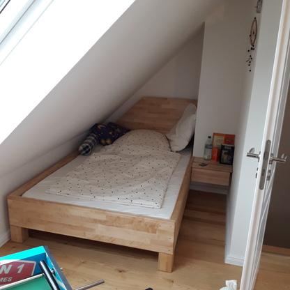 Holzbett unter Dachfenster Helles Holzbett steht passgenau unter einer Dachschräge neben einem kleinen Nachttisch.