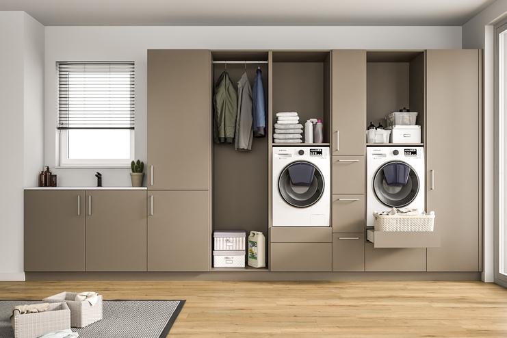 Modern gestalteter Wirtschaftsraum mit maßgefertigten Schränken. Ein heller Raum mit einem großen, maßgeschneiderten Schranksystem in Taupe, integrierten Waschmaschinen, Ablageflächen und einem Fenster mit Jalousien.